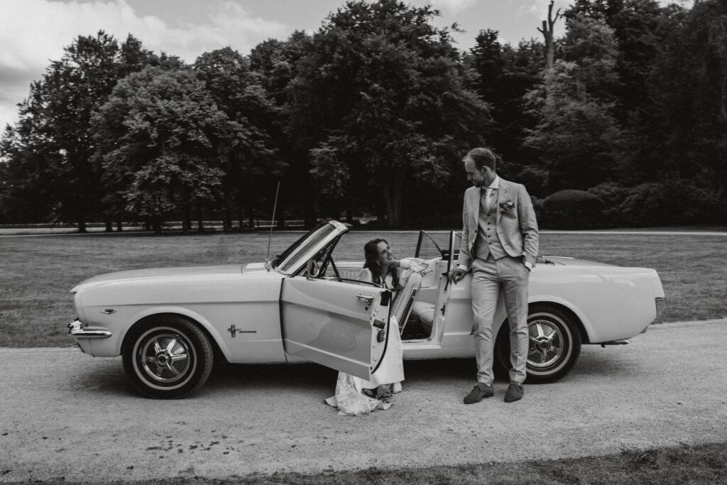 Bruid zit in bruidsauto en bruidegom leunt er tegenaan