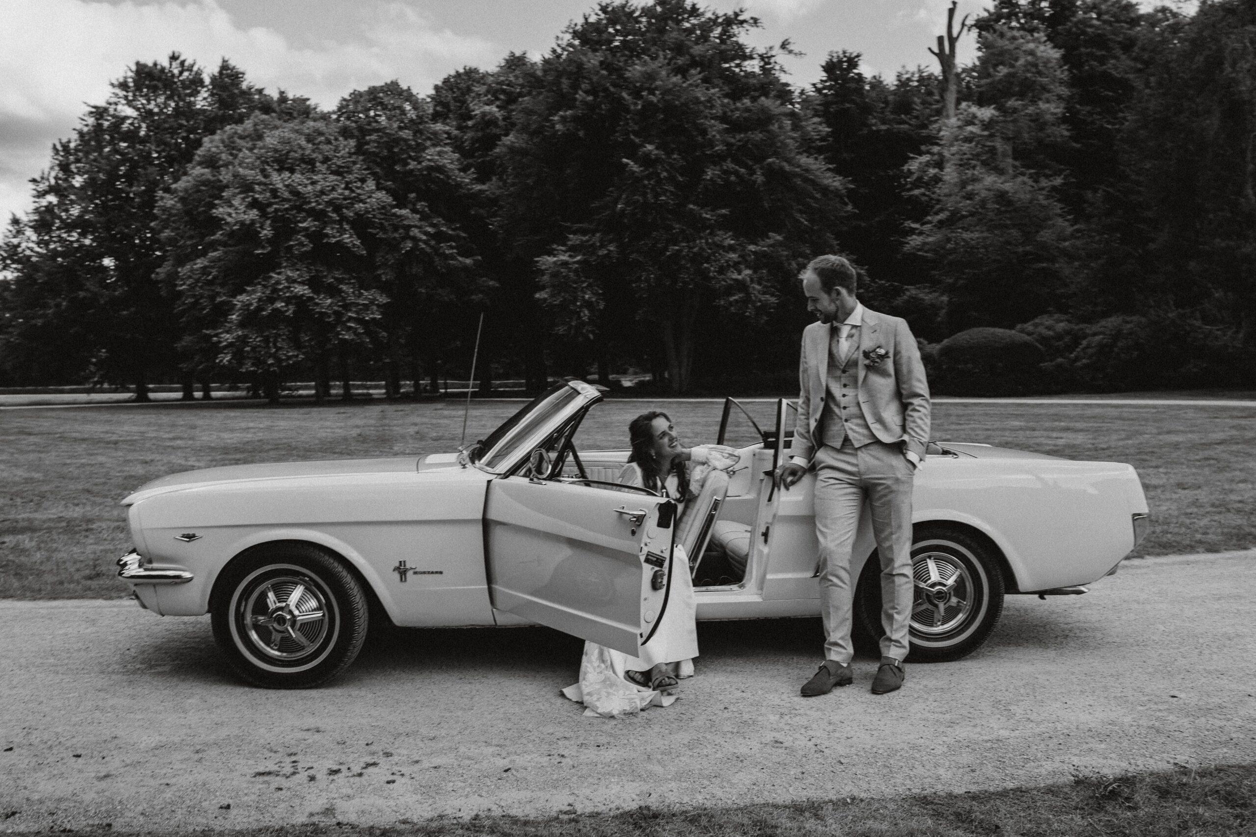 Bruid zit in bruidsauto en bruidegom leunt er tegenaan