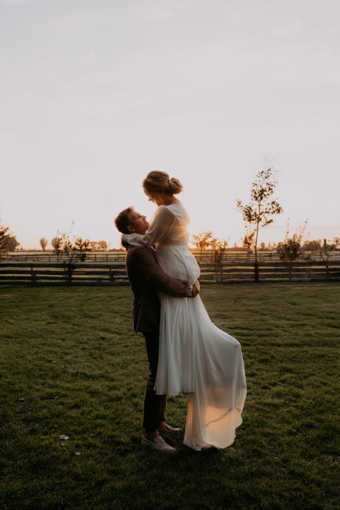 Bruidegom tilt bruid op in weiland tijdens golden hour
